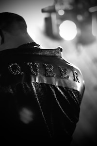 The image features a close-up view of a person from behind, wearing a jacket with the word 'QUEER' studded or embroidered on the back. The image is in black and white, with a focus on the texture of the jacket.