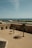 A coastal fortress with palm trees casting shadows across a large courtyard. The ocean is visible in the distance, along with a line of palm trees along a promenade. Three people are walking in the courtyard.