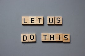 Wooden tiles with black letters spell out the motivational phrase 'LET US DO THIS' arranged on a gray background.