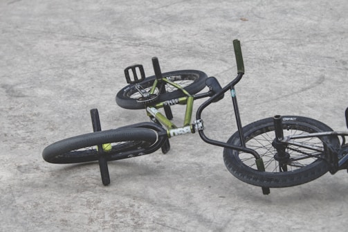 A BMX bike is lying on its side on a concrete surface. The black tires contrast with the green frame and handlebars. The setting appears to be an outdoor area, likely a skate park or similar environment.