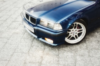 A close-up view of a blue car with a shiny metallic finish, highlighting the front bumper, headlight, and a polished alloy wheel. The car is parked on a light-gray, textured pavement.