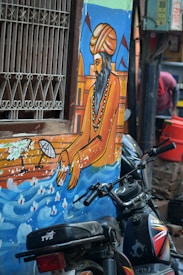 A vibrant mural is painted on the side of a building, depicting a man with a turban and beaded necklace, possibly of historical or cultural significance. He is portrayed with an outstretched hand reaching towards a body of water. The mural includes elements of traditional architecture and flags in the background. In front of the mural, there are motorcycles parked, partially obscuring the artwork.