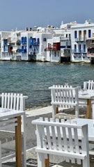 A picturesque waterside scene featuring traditional whitewashed buildings with vibrant blue and red accents. The calm sea flows gently against the backdrop of these buildings, while in the foreground, white wooden chairs and tables are arranged invitingly on a stone patio, suggesting a seaside dining area.