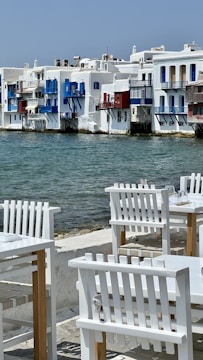 A picturesque waterside scene featuring traditional whitewashed buildings with vibrant blue and red accents. The calm sea flows gently against the backdrop of these buildings, while in the foreground, white wooden chairs and tables are arranged invitingly on a stone patio, suggesting a seaside dining area.