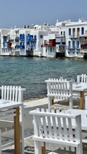 A picturesque waterside scene featuring traditional whitewashed buildings with vibrant blue and red accents. The calm sea flows gently against the backdrop of these buildings, while in the foreground, white wooden chairs and tables are arranged invitingly on a stone patio, suggesting a seaside dining area.