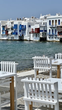 A picturesque waterside scene featuring traditional whitewashed buildings with vibrant blue and red accents. The calm sea flows gently against the backdrop of these buildings, while in the foreground, white wooden chairs and tables are arranged invitingly on a stone patio, suggesting a seaside dining area.