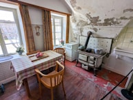 A rustic, vintage kitchen featuring a worn wooden floor and a small table covered with a checkered cloth. There are wooden chairs and a classic wood-burning stove with pots on top. The walls show signs of wear with peeling paint. Two large windows with partially opened curtains allow natural light to fill the room, and there are potted plants on the windowsills. A white, small sink is visible next to the stove.