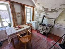 A rustic, vintage kitchen featuring a worn wooden floor and a small table covered with a checkered cloth. There are wooden chairs and a classic wood-burning stove with pots on top. The walls show signs of wear with peeling paint. Two large windows with partially opened curtains allow natural light to fill the room, and there are potted plants on the windowsills. A white, small sink is visible next to the stove.