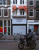 A street scene features a vintage-style movie theater facade with a large red sign reading 'THE MOVIES'. Below the sign, the marquee displays a message and a website. The building is bordered by traditional brick facades and has several bicycles parked at the front. A person dressed in dark clothing and riding a motorized scooter moves past the theater, creating a blurred motion effect.