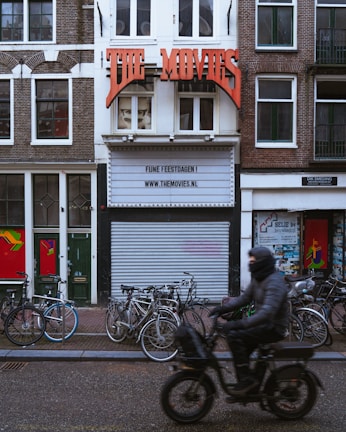 A street scene features a vintage-style movie theater facade with a large red sign reading 'THE MOVIES'. Below the sign, the marquee displays a message and a website. The building is bordered by traditional brick facades and has several bicycles parked at the front. A person dressed in dark clothing and riding a motorized scooter moves past the theater, creating a blurred motion effect.