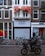 A street scene features a vintage-style movie theater facade with a large red sign reading 'THE MOVIES'. Below the sign, the marquee displays a message and a website. The building is bordered by traditional brick facades and has several bicycles parked at the front. A person dressed in dark clothing and riding a motorized scooter moves past the theater, creating a blurred motion effect.