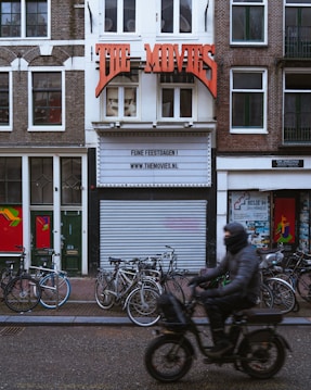 A street scene features a vintage-style movie theater facade with a large red sign reading 'THE MOVIES'. Below the sign, the marquee displays a message and a website. The building is bordered by traditional brick facades and has several bicycles parked at the front. A person dressed in dark clothing and riding a motorized scooter moves past the theater, creating a blurred motion effect.