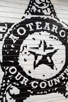 The image depicts a bold mural painted on a panel with large text and a central star emblem. The words 'Aotearoa' and 'Our Country' are prominently displayed in white against a black background, suggesting a strong cultural or national theme.