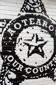 The image depicts a bold mural painted on a panel with large text and a central star emblem. The words 'Aotearoa' and 'Our Country' are prominently displayed in white against a black background, suggesting a strong cultural or national theme.