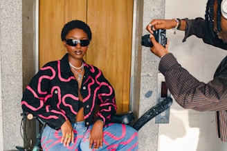 A stylish person wearing a dark jacket with pink patterns and sunglasses sits confidently in a chair. They are being photographed by another individual holding a camera. The setting appears to be indoors, with a wooden door and stone wall in the background.
