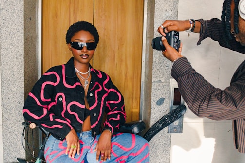 A stylish person wearing a dark jacket with pink patterns and sunglasses sits confidently in a chair. They are being photographed by another individual holding a camera. The setting appears to be indoors, with a wooden door and stone wall in the background.