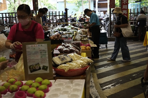 A bustling market scene with several people shopping for fresh produce and other goods. Various items are displayed on tables, including green apples, packaged foods, and a variety of vegetables. The market appears to be indoors or semi-outdoor with natural light coming through the openings.
