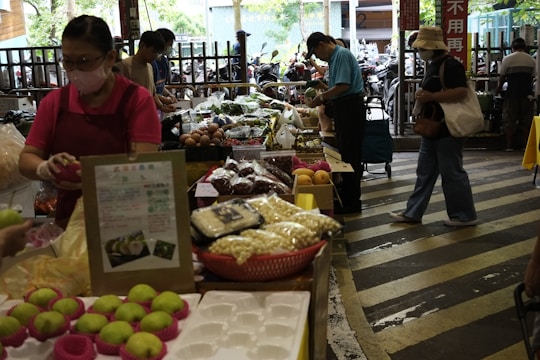 A bustling market scene with several people shopping for fresh produce and other goods. Various items are displayed on tables, including green apples, packaged foods, and a variety of vegetables. The market appears to be indoors or semi-outdoor with natural light coming through the openings.