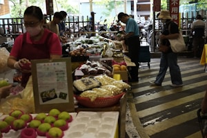 A bustling market scene with several people shopping for fresh produce and other goods. Various items are displayed on tables, including green apples, packaged foods, and a variety of vegetables. The market appears to be indoors or semi-outdoor with natural light coming through the openings.