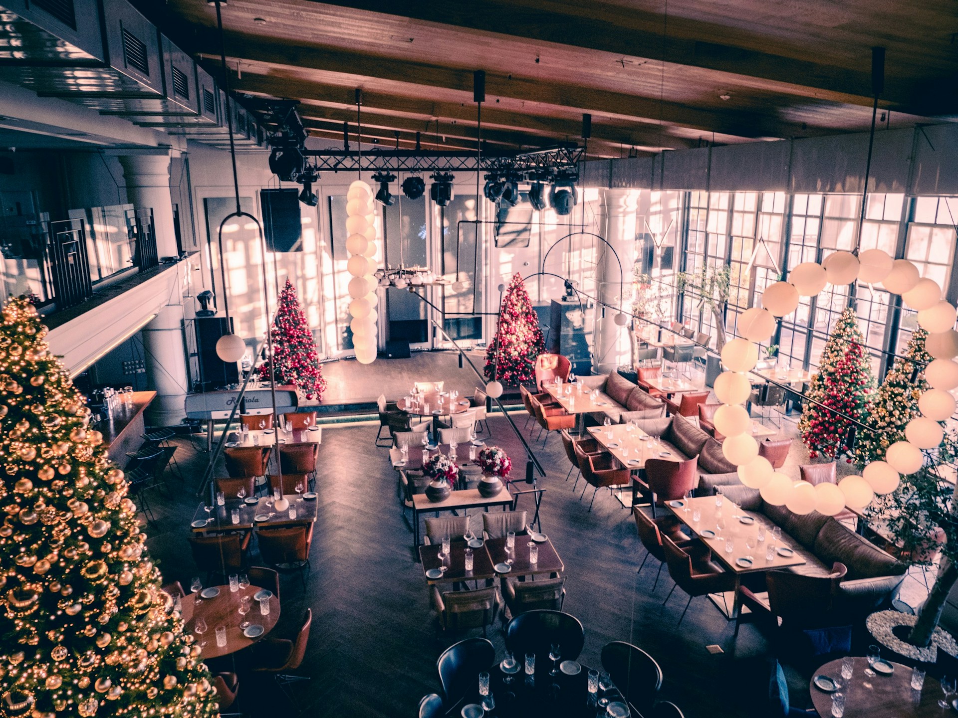 A warmly decorated dining area featuring several Christmas trees adorned with lights and ornaments. The space has a high, wooden ceiling and large windows allowing natural light to fill the room. Tables are set with glasses and cutlery, while comfortable seating arrangements include sofas and chairs. Decorative light fixtures hang from the ceiling, enhancing the festive atmosphere.