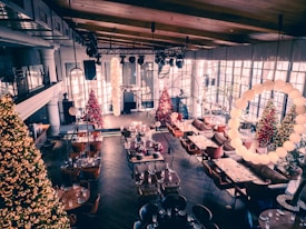 A warmly decorated dining area featuring several Christmas trees adorned with lights and ornaments. The space has a high, wooden ceiling and large windows allowing natural light to fill the room. Tables are set with glasses and cutlery, while comfortable seating arrangements include sofas and chairs. Decorative light fixtures hang from the ceiling, enhancing the festive atmosphere.