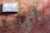 A tiled street sign with blue decorative elements and the text 'Calle de los Suspiros' is affixed to an old, weather-worn wall. The wall is characterized by its rough texture and a mix of faded colors, including red, brown, and patches of green moss or lichen.
