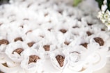 A collection of chocolate confections wrapped in delicate, lacy white paper is arranged on an ornate, white serving tray. The chocolates are topped with small, crunchy pieces and are evenly spaced, creating an aesthetically pleasing pattern. Soft natural lighting adds an elegant touch to the overall presentation.