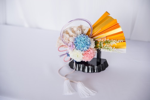 A decorative arrangement features a collection of pastel-colored flowers, including blue, pink, and white blooms. It is accented with a golden folding fan and delicate pink and purple ribbons. The entire composition is set on a black stand with a textured base, and a white tassel cord is draped elegantly to the side.