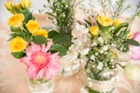 A collection of colorful flowers in glass jars, featuring vibrant yellow roses and a prominent pink gerbera daisy. Additional greenery and small white flowers add texture and a natural feel to the arrangement.