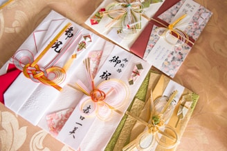 Intricately designed Japanese envelopes used for gifting, adorned with decorative knots made from colorful cords and accented with traditional paper and floral patterns. The envelopes display elegant calligraphy and are arranged on a patterned fabric background.