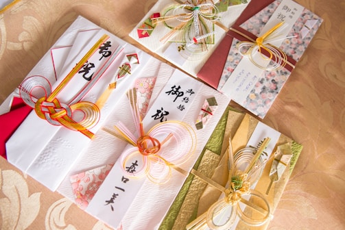 Intricately designed Japanese envelopes used for gifting, adorned with decorative knots made from colorful cords and accented with traditional paper and floral patterns. The envelopes display elegant calligraphy and are arranged on a patterned fabric background.