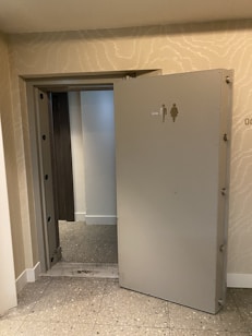 An open heavy door, resembling a vault or secure access point, leads into a small, tiled area. The walls are a light beige color with subtle wave-like patterns. On the door is a sign depicting male and female figures with an arrow indicating direction.