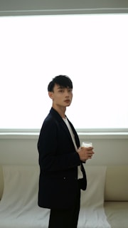 A person wearing a dark blazer stands holding a drink against a backdrop with soft natural lighting. The room appears minimalistic with a neutral-colored sofa and a large window letting in bright light.