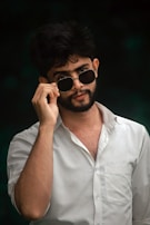 A man is wearing a white shirt and adjusting his round sunglasses, with a confident expression. His dark hair is slightly tousled and there is a dark background that enhances the contrast.
