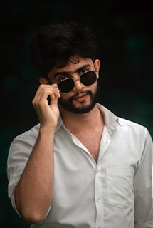 A man is wearing a white shirt and adjusting his round sunglasses, with a confident expression. His dark hair is slightly tousled and there is a dark background that enhances the contrast.