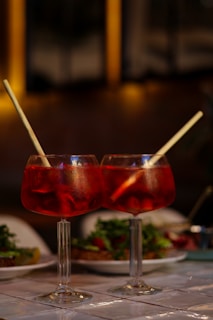 Two large red cocktails with ice and straws sit on a table, with plates of food and a blurred background creating a warm and inviting atmosphere.