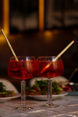 Two large red cocktails with ice and straws sit on a table, with plates of food and a blurred background creating a warm and inviting atmosphere.