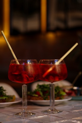 Two large red cocktails with ice and straws sit on a table, with plates of food and a blurred background creating a warm and inviting atmosphere.