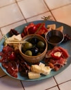 A charcuterie board featuring a variety of cured meats, cheeses, and olives is arranged on a teal plate. A small dish in the center holds green and black olives. The board is garnished with almonds and stuffed red peppers, with skewers for serving. The setup is on a tiled surface, giving a casual yet elegant dining vibe.