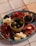 A charcuterie board featuring a variety of cured meats, cheeses, and olives is arranged on a teal plate. A small dish in the center holds green and black olives. The board is garnished with almonds and stuffed red peppers, with skewers for serving. The setup is on a tiled surface, giving a casual yet elegant dining vibe.