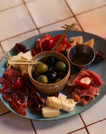 A charcuterie board featuring a variety of cured meats, cheeses, and olives is arranged on a teal plate. A small dish in the center holds green and black olives. The board is garnished with almonds and stuffed red peppers, with skewers for serving. The setup is on a tiled surface, giving a casual yet elegant dining vibe.