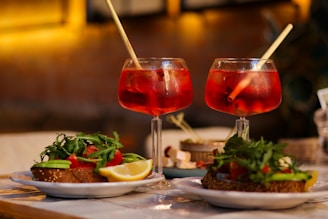 Two glasses of red cocktails each with a straw rest on a table. In the background, the image is blurred, suggesting a cozy indoor setting, possibly a restaurant or cafe. In the foreground, there are plates of food, including sandwiches topped with greens, cherry tomatoes, and lemon wedges.