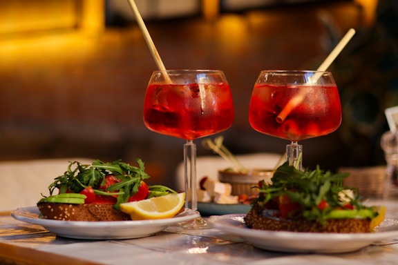Two glasses of red cocktails each with a straw rest on a table. In the background, the image is blurred, suggesting a cozy indoor setting, possibly a restaurant or cafe. In the foreground, there are plates of food, including sandwiches topped with greens, cherry tomatoes, and lemon wedges.