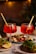 Two elegant stemmed glasses filled with a vibrant red beverage, topped with ice cubes and garnished with a straw. The glasses are placed on a table next to plates of appetizers, featuring slices of bread topped with arugula, tomatoes, and lemon wedges. In the background, small dishes contain a variety of cheeses and cured meats.