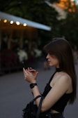 A young woman is standing outdoors, focused intently on her smartphone. She is wearing a black sleeveless top and a smartwatch on her wrist, and her long brown hair is partially covering her face. The background shows defocused lights and greenery, suggesting an evening or late afternoon setting in a park or public area.