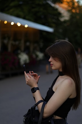A young woman is standing outdoors, focused intently on her smartphone. She is wearing a black sleeveless top and a smartwatch on her wrist, and her long brown hair is partially covering her face. The background shows defocused lights and greenery, suggesting an evening or late afternoon setting in a park or public area.