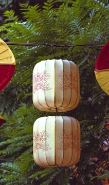 Two intricately designed lanterns are suspended in front of lush green foliage. The lanterns are cylindrical with a cream color and red decorative patterns. They are connected by a central rod, which is part of a larger hanging structure. The background consists of leafy branches, adding a natural contrast to the artificial lanterns.