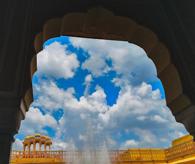 An architectural arch frames a view of a vibrant blue sky filled with fluffy white clouds. Below, a section of ornate yellow building with intricate designs is visible, hinting at traditional architecture. A water fountain can be seen in action at the bottom of the image.