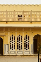 A detailed architectural facade featuring a series of tall, arched windows with intricate geometric patterns. The structure is adorned with ornate grillwork, and the walls are painted in warm shades of yellow and cream. Classical elements are visible, such as carved borders and decorative motifs.