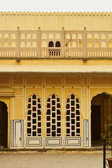 A detailed architectural facade featuring a series of tall, arched windows with intricate geometric patterns. The structure is adorned with ornate grillwork, and the walls are painted in warm shades of yellow and cream. Classical elements are visible, such as carved borders and decorative motifs.